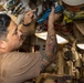 A tattooed male technician or maintenance worker is working on overhead piping and valve systems in what appears to be a ship's engine room or mechanical compartment. He is wearing a tan/brown t-shirt and a wristwatch, and is manipulating blue-handled valves on a complex pipe assembly. The environment is a narrow industrial corridor with multiple pipes, fittings, and mechanical components visible.