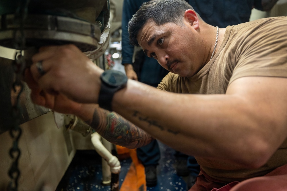A man in a tan/olive t-shirt is performing mechanical or maintenance work, reaching into a tight space with both hands. He appears focused and concentrated on the task, working in what appears to be an industrial or military vessel environment. A second person is partially visible in the background.