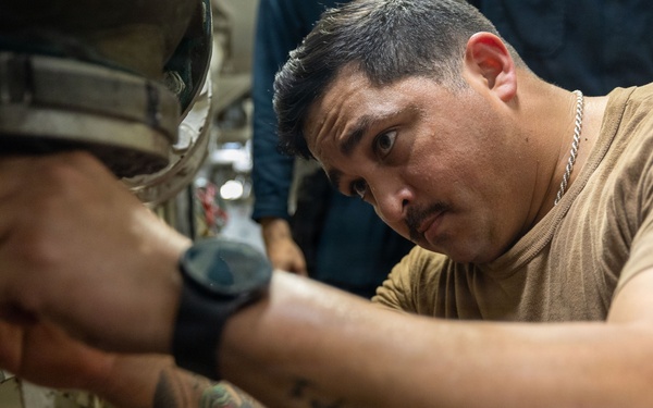 A man in a tan/olive t-shirt is performing mechanical or maintenance work, reaching into a tight space with both hands. He appears focused and concentrated on the task, working in what appears to be an industrial or military vessel environment. A second person is partially visible in the background.