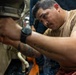 A man in a tan/olive t-shirt is performing mechanical or maintenance work, reaching into a tight space with both hands. He appears focused and concentrated on the task, working in what appears to be an industrial or military vessel environment. A second person is partially visible in the background.