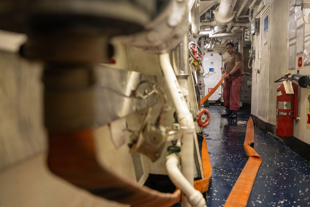 A crew member is working in a narrow interior passageway of what appears to be a naval vessel or large ship. The person is handling an orange hose or strap in a confined corridor with exposed piping, machinery, and safety equipment. The wet floor and industrial environment suggest ongoing maintenance or damage control operations.