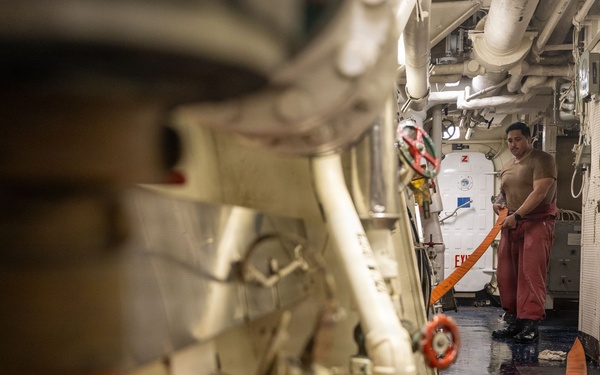A crew member is working in a narrow interior passageway of what appears to be a naval vessel or large ship. The person is handling an orange hose or strap in a confined corridor with exposed piping, machinery, and safety equipment. The wet floor and industrial environment suggest ongoing maintenance or damage control operations.