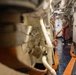 A crew member is working in a narrow interior passageway of what appears to be a naval vessel or large ship. The person is handling an orange hose or strap in a confined corridor with exposed piping, machinery, and safety equipment. The wet floor and industrial environment suggest ongoing maintenance or damage control operations.