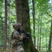 A U.S. Army soldier in full combat gear is kneeling behind a large moss-covered tree in a dense forest, aiming a rifle with an optic scope. The soldier is wearing Operational Camouflage Pattern (OCP) uniform with a camouflaged helmet, tactical vest, and full kit. The setting appears to be a training exercise in a European-style deciduous/mixed forest.