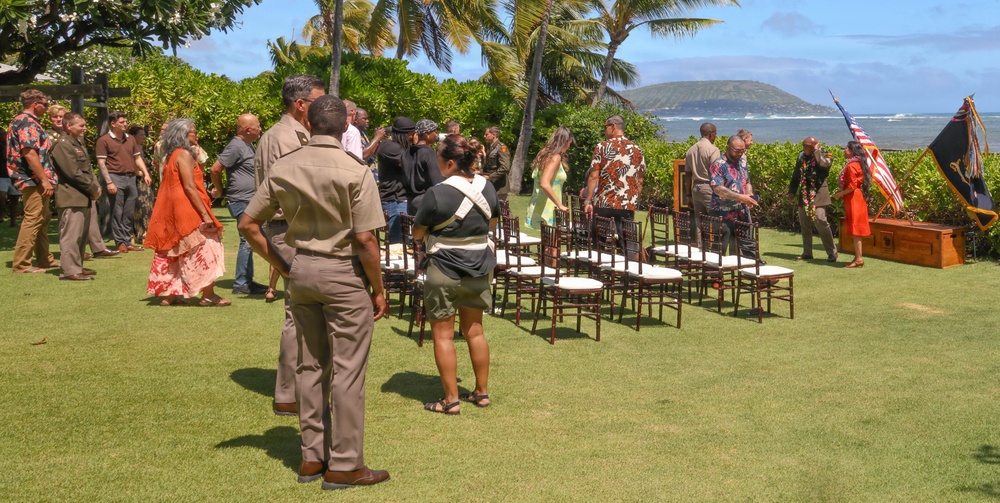 An outdoor ceremony or gathering is taking place on a well-manicured lawn in what appears to be Hawaii, identifiable by the palm trees, ocean backdrop, and mountainous island in the distance. Several individuals in military uniforms (tan/khaki) are present alongside civilians in casual and semi-formal attire. Rows of wooden chairs are arranged on the grass, and an American flag along with another flag on a stand are visible on the right side.