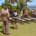 An outdoor ceremony or gathering is taking place on a well-manicured lawn in what appears to be Hawaii, identifiable by the palm trees, ocean backdrop, and mountainous island in the distance. Several individuals in military uniforms (tan/khaki) are present alongside civilians in casual and semi-formal attire. Rows of wooden chairs are arranged on the grass, and an American flag along with another flag on a stand are visible on the right side.