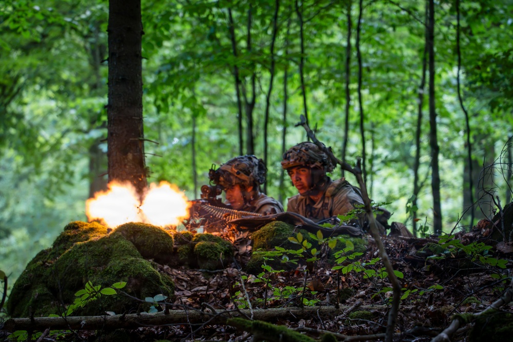 Two soldiers in camouflage uniforms and helmets are positioned behind a moss-covered earthen berm in a dense deciduous forest. A machine gun is being fired, producing a visible muzzle flash and what appears to be a belt-fed ammunition strip. The scene appears to be a military training exercise or combat operation in a woodland environment.