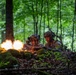 Two soldiers in camouflage uniforms and helmets are positioned behind a moss-covered earthen berm in a dense deciduous forest. A machine gun is being fired, producing a visible muzzle flash and what appears to be a belt-fed ammunition strip. The scene appears to be a military training exercise or combat operation in a woodland environment.
