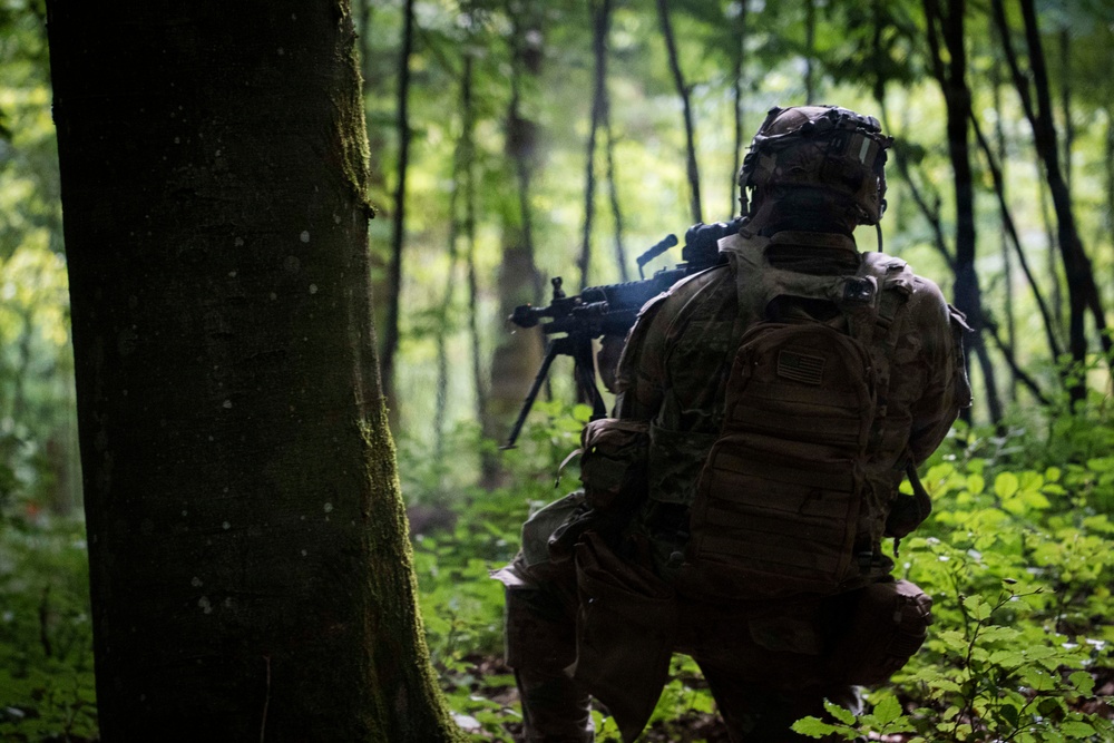 A soldier in full combat gear is kneeling in a forested environment, facing away from the camera toward a deployed weapon system on a bipod. The soldier is wearing MultiCam/OCP camouflage pattern with a tactical backpack bearing a visible U.S. flag patch. The scene appears to be a military training exercise or operational patrol in a dense, green woodland setting.