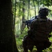 A soldier in full combat gear is kneeling in a forested environment, facing away from the camera toward a deployed weapon system on a bipod. The soldier is wearing MultiCam/OCP camouflage pattern with a tactical backpack bearing a visible U.S. flag patch. The scene appears to be a military training exercise or operational patrol in a dense, green woodland setting.