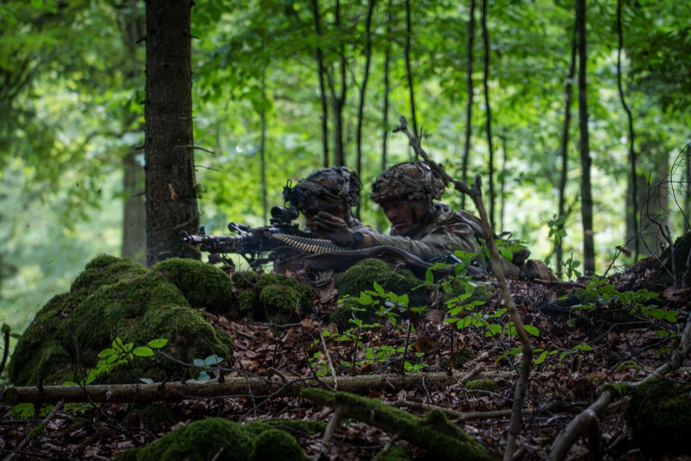 Two soldiers in full camouflage gear are positioned behind a moss-covered natural berm in a dense deciduous forest. They appear to be in a defensive firing position, with one soldier aiming a belt-fed machine gun forward. The lush green foliage and leaf-covered forest floor suggest a temperate woodland environment during summer or early autumn.