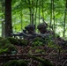 Two soldiers in full camouflage gear are positioned behind a moss-covered natural berm in a dense deciduous forest. They appear to be in a defensive firing position, with one soldier aiming a belt-fed machine gun forward. The lush green foliage and leaf-covered forest floor suggest a temperate woodland environment during summer or early autumn.