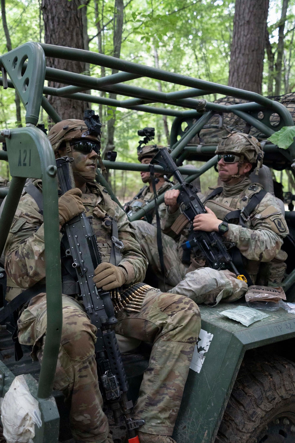 Multiple soldiers in full combat gear are seated in a military utility vehicle (likely an LMTV or similar tactical vehicle) in a wooded training environment. The soldiers are wearing MultiCam/OCP camouflage uniforms with face paint, helmets equipped with night vision mounts, and tactical sunglasses. The scene appears to be a military field training exercise based on the equipment, vehicle markings, and forest setting.