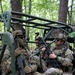 Multiple soldiers in full combat gear are seated in a military utility vehicle (likely an LMTV or similar tactical vehicle) in a wooded training environment. The soldiers are wearing MultiCam/OCP camouflage uniforms with face paint, helmets equipped with night vision mounts, and tactical sunglasses. The scene appears to be a military field training exercise based on the equipment, vehicle markings, and forest setting.
