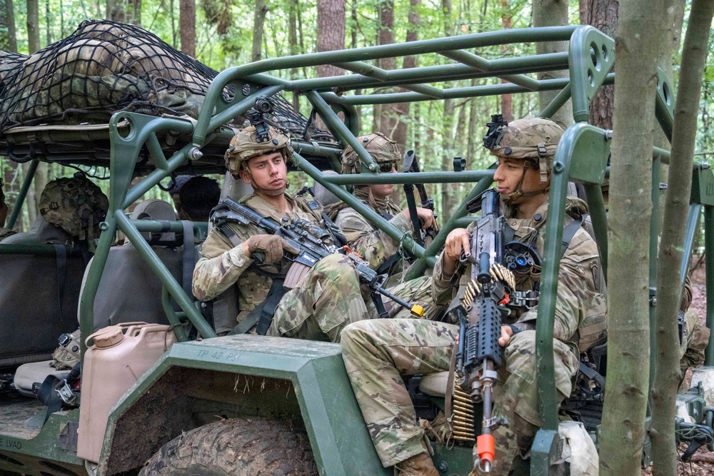 Multiple soldiers in US Army OCP (Operational Camouflage Pattern) uniforms are seated in a military utility vehicle, likely an Polaris MRZR or similar light tactical vehicle, in a forested environment. The soldiers are armed and equipped with combat gear including helmets with night vision mounts, body armor, and weapons including what appears to be an M4 carbine and an M249 SAW (Squad Automatic Weapon) with a linked ammunition belt. The scene appears to be a training exercise or tactical operation pause in a wooded area.