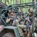 Multiple soldiers in US Army OCP (Operational Camouflage Pattern) uniforms are seated in a military utility vehicle, likely an Polaris MRZR or similar light tactical vehicle, in a forested environment. The soldiers are armed and equipped with combat gear including helmets with night vision mounts, body armor, and weapons including what appears to be an M4 carbine and an M249 SAW (Squad Automatic Weapon) with a linked ammunition belt. The scene appears to be a training exercise or tactical operation pause in a wooded area.