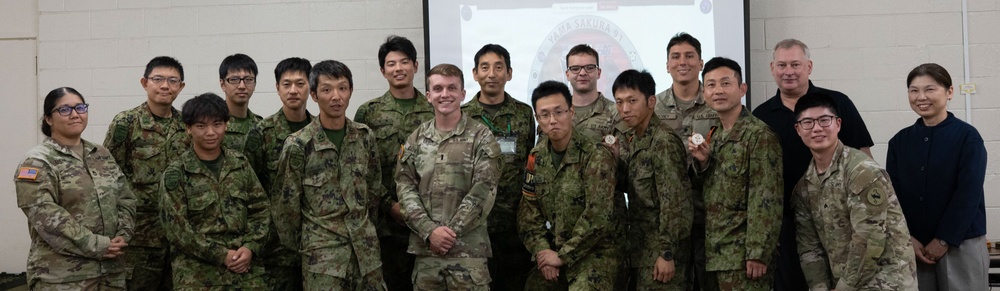 A group of approximately 16 military personnel and two civilians pose for a formal group photo in front of a projection screen. The group includes soldiers wearing U.S. Army camouflage uniforms (OCP pattern) and what appear to be Japanese Ground Self-Defense Force uniforms, suggesting a joint military exercise or event. The projection screen behind them displays what appears to be a 'Yama Sakura' exercise logo, indicating this is likely a U.S.-Japan bilateral military exercise.