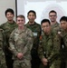 A group of approximately 16 military personnel and two civilians pose for a formal group photo in front of a projection screen. The group includes soldiers wearing U.S. Army camouflage uniforms (OCP pattern) and what appear to be Japanese Ground Self-Defense Force uniforms, suggesting a joint military exercise or event. The projection screen behind them displays what appears to be a 'Yama Sakura' exercise logo, indicating this is likely a U.S.-Japan bilateral military exercise.