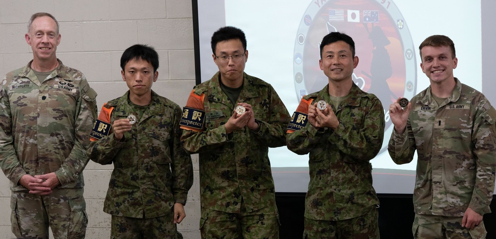 Five military personnel pose together for a photo, with three Japanese Ground Self-Defense Force soldiers wearing 'Interpreter' armbands in both English and Japanese kanji. Four of the five individuals are holding challenge coins or medallions. The setting appears to be an indoor military briefing room with a projected emblem showing US, Japanese, and Australian flags, suggesting a multinational military exercise or event.