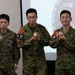 Five military personnel pose together for a photo, with three Japanese Ground Self-Defense Force soldiers wearing 'Interpreter' armbands in both English and Japanese kanji. Four of the five individuals are holding challenge coins or medallions. The setting appears to be an indoor military briefing room with a projected emblem showing US, Japanese, and Australian flags, suggesting a multinational military exercise or event.