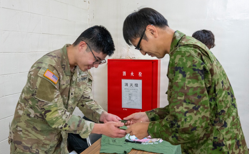 A U.S. Army soldier and a Japan Ground Self-Defense Force (JGSDF) soldier are engaged in a collaborative activity at a table, appearing to exchange or sign items such as t-shirts. The setting appears to be an indoor facility in Japan, evidenced by the red fire hydrant cabinet on the wall with Japanese characters reading '消火栓' (fire hydrant/hose cabinet). This appears to be a bilateral military exchange or goodwill event.