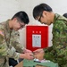 A U.S. Army soldier and a Japan Ground Self-Defense Force (JGSDF) soldier are engaged in a collaborative activity at a table, appearing to exchange or sign items such as t-shirts. The setting appears to be an indoor facility in Japan, evidenced by the red fire hydrant cabinet on the wall with Japanese characters reading '消火栓' (fire hydrant/hose cabinet). This appears to be a bilateral military exchange or goodwill event.