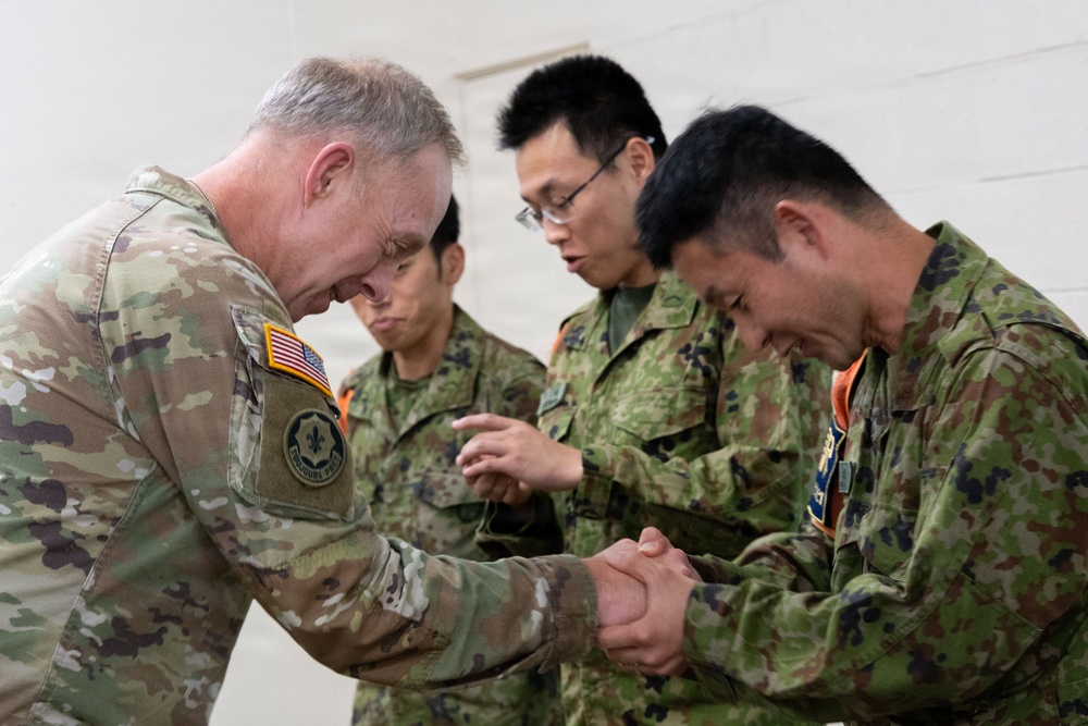 A U.S. Army soldier in OCP (Operational Camouflage Pattern) uniform is exchanging a coin or small item with a member of the Japan Ground Self-Defense Force (JGSDF), in what appears to be a formal military coin ceremony. Three JGSDF soldiers in their distinctive pixelated camouflage uniforms are present, with two bowing slightly in a gesture of respect. The setting suggests a bilateral military exchange or joint training event.