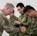 A U.S. Army soldier in OCP (Operational Camouflage Pattern) uniform is exchanging a coin or small item with a member of the Japan Ground Self-Defense Force (JGSDF), in what appears to be a formal military coin ceremony. Three JGSDF soldiers in their distinctive pixelated camouflage uniforms are present, with two bowing slightly in a gesture of respect. The setting suggests a bilateral military exchange or joint training event.