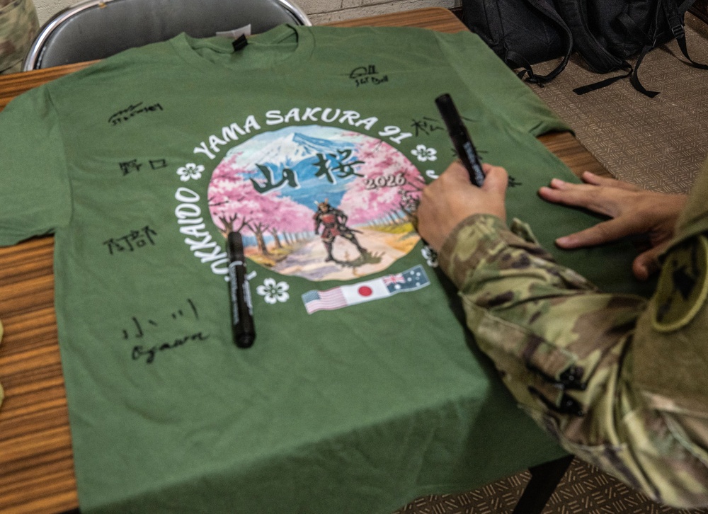 A green t-shirt commemorating 'Yama Sakura 91' military exercise is laid flat on a wooden table and is being signed with markers. The shirt features a circular graphic design with cherry blossoms, a samurai figure, Mount Fuji, and the flags of the United States, Japan, and Australia, along with the year 2026 and the location 'Hokkaido'. Multiple signatures in both English and Japanese characters are already visible on the shirt.