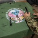 A green t-shirt commemorating 'Yama Sakura 91' military exercise is laid flat on a wooden table and is being signed with markers. The shirt features a circular graphic design with cherry blossoms, a samurai figure, Mount Fuji, and the flags of the United States, Japan, and Australia, along with the year 2026 and the location 'Hokkaido'. Multiple signatures in both English and Japanese characters are already visible on the shirt.