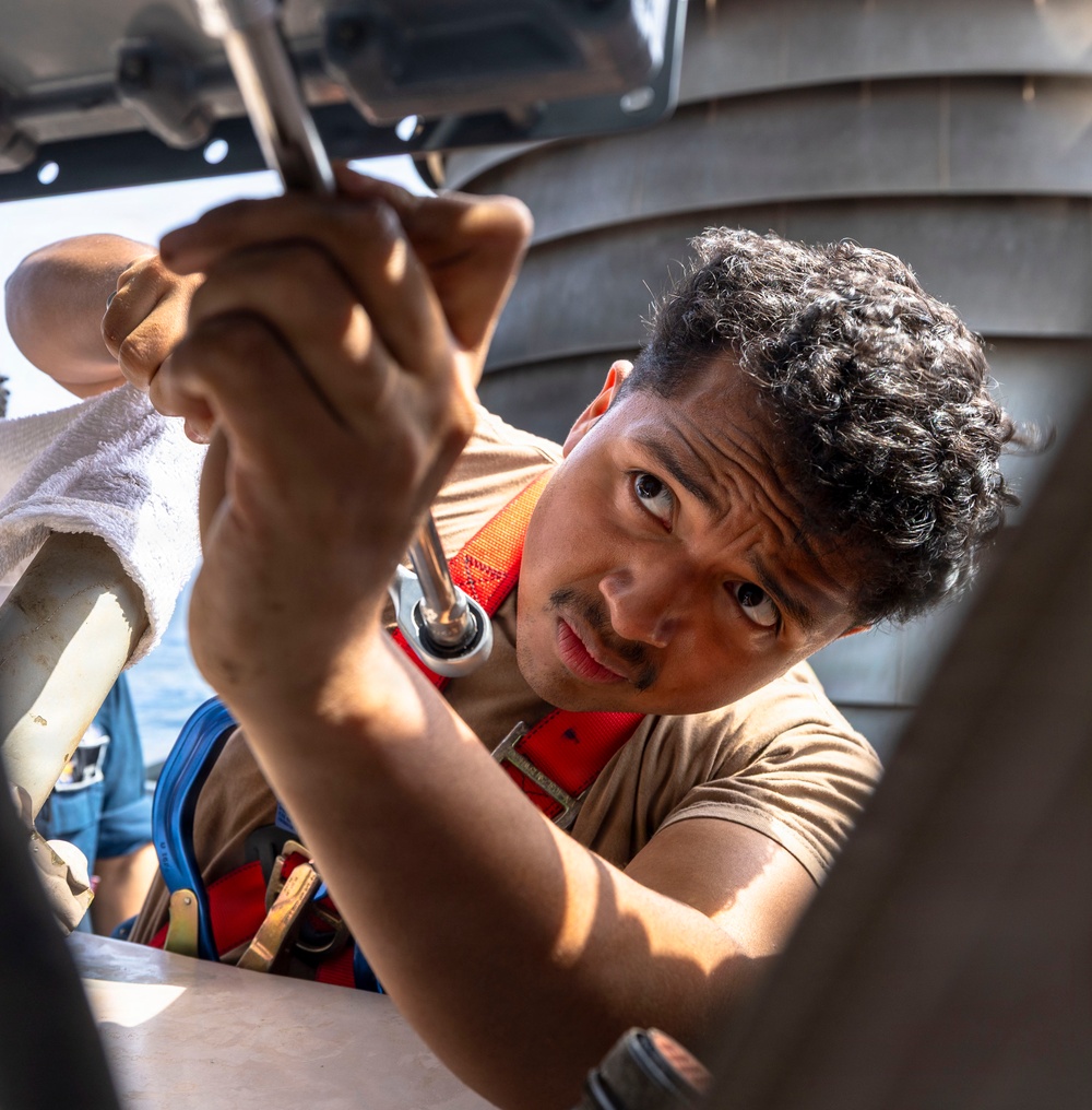 A young man wearing a tan shirt and an orange safety harness is using a wrench or ratchet tool to perform mechanical maintenance work on what appears to be a naval vessel or boat. He is working in a confined or overhead space, concentrating intently on the task. The outdoor maritime setting is visible in the background with water and vessel structures.