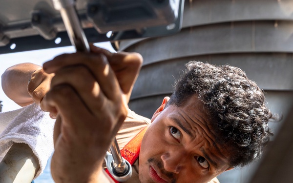 A young man wearing a tan shirt and an orange safety harness is using a wrench or ratchet tool to perform mechanical maintenance work on what appears to be a naval vessel or boat. He is working in a confined or overhead space, concentrating intently on the task. The outdoor maritime setting is visible in the background with water and vessel structures.