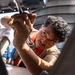 A young man wearing a tan shirt and an orange safety harness is using a wrench or ratchet tool to perform mechanical maintenance work on what appears to be a naval vessel or boat. He is working in a confined or overhead space, concentrating intently on the task. The outdoor maritime setting is visible in the background with water and vessel structures.