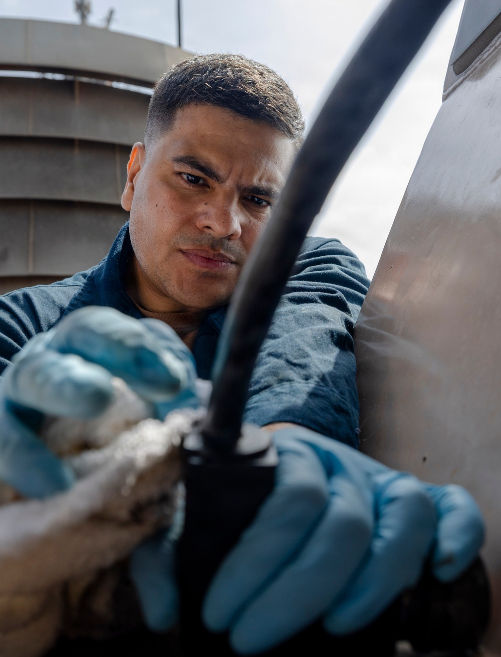A man wearing blue work gloves is performing a maintenance or repair task on what appears to be a large metal surface or vehicle exterior. He is focused intently on manipulating a black cable or hose connection with both hands. The image is taken from a low angle, with an industrial structure visible in the background.