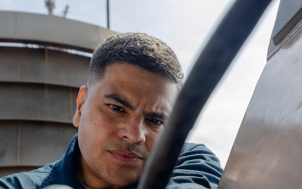 A man wearing blue work gloves is performing a maintenance or repair task on what appears to be a large metal surface or vehicle exterior. He is focused intently on manipulating a black cable or hose connection with both hands. The image is taken from a low angle, with an industrial structure visible in the background.