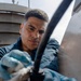 A man wearing blue work gloves is performing a maintenance or repair task on what appears to be a large metal surface or vehicle exterior. He is focused intently on manipulating a black cable or hose connection with both hands. The image is taken from a low angle, with an industrial structure visible in the background.