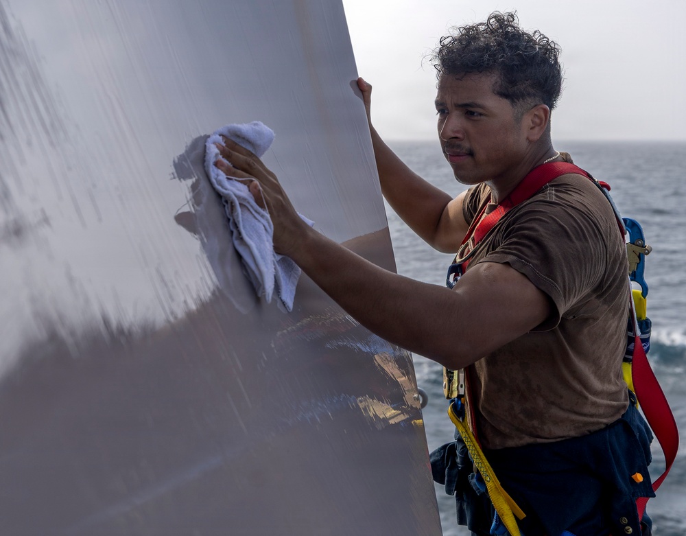 A young man wearing a safety harness is cleaning or polishing a large metallic surface, likely part of a naval vessel or ship structure. He is using a white cloth/rag to wipe the reflective gray surface while standing at an elevated or exposed position with the ocean visible in the background. The safety harness with red and yellow straps suggests this is an occupational task performed at height or in a potentially hazardous location.