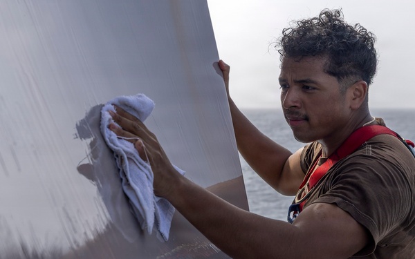 A young man wearing a safety harness is cleaning or polishing a large metallic surface, likely part of a naval vessel or ship structure. He is using a white cloth/rag to wipe the reflective gray surface while standing at an elevated or exposed position with the ocean visible in the background. The safety harness with red and yellow straps suggests this is an occupational task performed at height or in a potentially hazardous location.