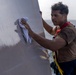 A young man wearing a safety harness is cleaning or polishing a large metallic surface, likely part of a naval vessel or ship structure. He is using a white cloth/rag to wipe the reflective gray surface while standing at an elevated or exposed position with the ocean visible in the background. The safety harness with red and yellow straps suggests this is an occupational task performed at height or in a potentially hazardous location.