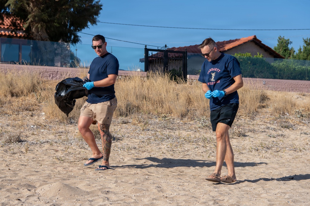 Two men wearing navy blue t-shirts and blue latex gloves are participating in a beach cleanup activity on a sandy area. The man on the left is carrying a filled black garbage bag while both scan the ground for litter. The setting appears to be a coastal or sandy area in a Mediterranean-style location, evidenced by the terracotta-roofed buildings in the background.