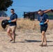 Two men wearing navy blue t-shirts and blue latex gloves are participating in a beach cleanup activity on a sandy area. The man on the left is carrying a filled black garbage bag while both scan the ground for litter. The setting appears to be a coastal or sandy area in a Mediterranean-style location, evidenced by the terracotta-roofed buildings in the background.