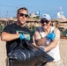 Two men are posing on a beach while holding a large black garbage bag, wearing blue latex gloves, suggesting they are participating in a beach cleanup activity. Both are wearing sunglasses and casual summer attire, smiling at the camera. The beach behind them features rows of lounge chairs with thatched umbrellas and a Blue Flag is visible in the background.