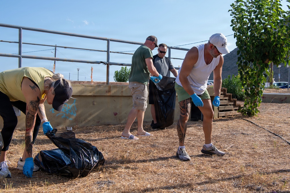 Four adults are engaged in an outdoor cleanup activity, collecting debris into large black garbage bags on a dry, grassy area. All participants are wearing blue protective gloves, suggesting an organized volunteer or community service effort. The setting appears to be a semi-urban outdoor space with raised garden beds, metal railings, and a mountainous background.
