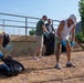 Four adults are engaged in an outdoor cleanup activity, collecting debris into large black garbage bags on a dry, grassy area. All participants are wearing blue protective gloves, suggesting an organized volunteer or community service effort. The setting appears to be a semi-urban outdoor space with raised garden beds, metal railings, and a mountainous background.