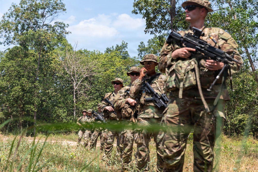 A column of military personnel in camouflage uniforms are lined up in a wooded, open terrain area, each carrying assault rifles. The soldiers appear to be conducting a patrol or field training exercise, standing in a single-file formation along a dirt path bordered by vegetation. The image is taken from a low angle, with the foreground soldier in sharp focus and the line receding into the background.