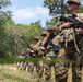 A column of military personnel in camouflage uniforms are lined up in a wooded, open terrain area, each carrying assault rifles. The soldiers appear to be conducting a patrol or field training exercise, standing in a single-file formation along a dirt path bordered by vegetation. The image is taken from a low angle, with the foreground soldier in sharp focus and the line receding into the background.