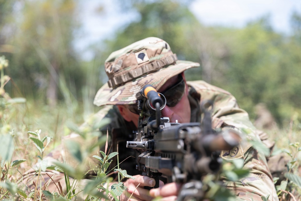 A soldier in multicam camouflage uniform and boonie hat is in a prone firing position in a field with low vegetation. The individual is aiming a machine gun or light support weapon equipped with an optical sight toward the camera. The setting appears to be an outdoor training or operational environment with trees visible in the blurred background.