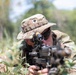 A soldier in multicam camouflage uniform and boonie hat is in a prone firing position in a field with low vegetation. The individual is aiming a machine gun or light support weapon equipped with an optical sight toward the camera. The setting appears to be an outdoor training or operational environment with trees visible in the blurred background.