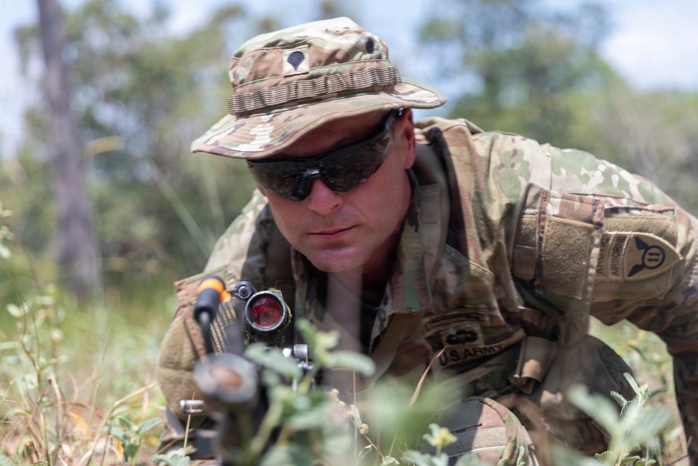 A U.S. Army soldier in full MultiCam/OCP camouflage uniform is positioned low in vegetation, aiming or observing through a weapon-mounted optic scope. The soldier wears a boonie hat, tactical sunglasses, body armor, and displays unit patches including what appears to be an Airborne or corps-level patch on the left shoulder. The image appears to be taken during a field training exercise or operational deployment.