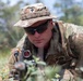 A U.S. Army soldier in full MultiCam/OCP camouflage uniform is positioned low in vegetation, aiming or observing through a weapon-mounted optic scope. The soldier wears a boonie hat, tactical sunglasses, body armor, and displays unit patches including what appears to be an Airborne or corps-level patch on the left shoulder. The image appears to be taken during a field training exercise or operational deployment.